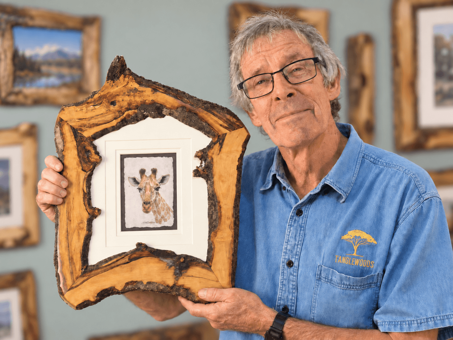 Brad Sharp holding a live-edge rustic frame with a giraffe drawing, in front of a wall of his frames